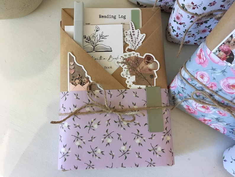 May include: A book wrapped in a light purple paper with a floral pattern. The book is tied with twine and has a brown paper envelope on top with a sticker that says "Reading Log".