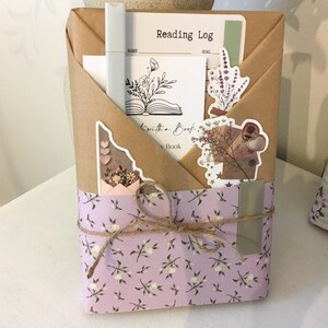 May include: A wrapped book with a purple and white floral design. The book is tied with twine and has a metal bookmark. The book is wrapped in brown paper with a sticker that says "Reading Log" and another sticker that says "Lost in a Book".