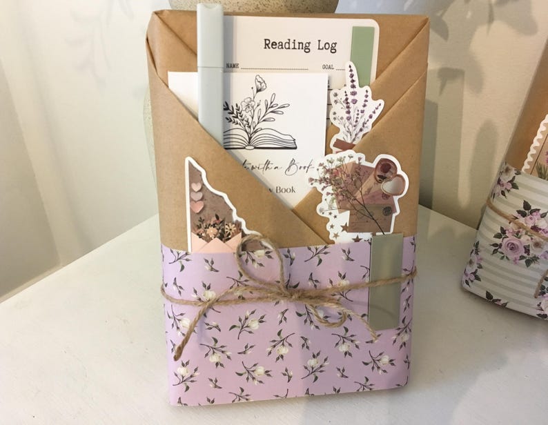 May include: A wrapped book with a purple and white floral design. The book is tied with twine and has a metal bookmark. The book is wrapped in brown paper with a sticker that says "Reading Log" and another sticker that says "Lost in a Book".