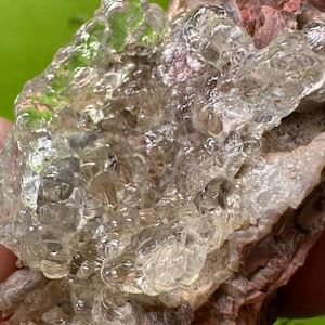 Puede incluir: Primer plano de un espécimen mineral natural. La piedra presenta una mezcla de formaciones cristalinas claras y roca de color marrón rojizo. La textura es desigual, con un aspecto burbujeante, casi líquido en algunas zonas.