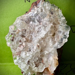 Puede incluir: Un espécimen mineral translúcido y cristalino con una superficie burbujeante y texturizada. El mineral tiene una apariencia clara y vítrea con toques de marrón y blanco. La forma general es irregular y orgánica.