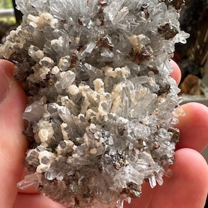 Può includere: Un ammasso di cristalli di quarzo trasparenti con un minerale metallico grigio argenteo. I cristalli sono appuntiti e acuminati, e il minerale metallico è disperso in tutto l'ammasso.
