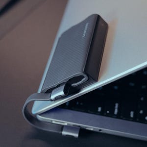 May include: A dark gray Samsung external hard drive connected to a silver laptop. The hard drive has a textured surface and a silver cable. The laptop is open, revealing the keyboard.