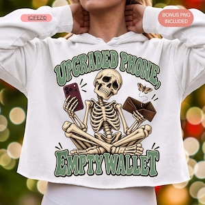 Könnte beinhalten: Ein weißer Kapuzenpullover mit einer Grafik eines im Schneidersitz sitzenden Skeletts, das ein dunkelrotes Smartphone in der einen und eine geöffnete braune Brieftasche in der anderen Hand hält. Der Text UPGRADED PHONE, EMPTY WALLET ist abgebildet.