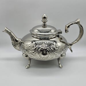 Op de afbeelding: Een zilverkleurige theepot met sierlijke bloemen- en krulversieringen. De theepot heeft een gebogen handvat, een tuit, een deksel met een decoratieve knop en staat op drie kleine pootjes. De theepot is waarschijnlijk van metaal.