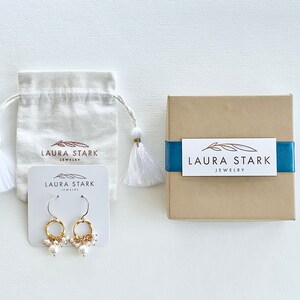 Pu&ograve; includere: Un paio di orecchini a cerchio dorati con piccole perle bianche che pendono dal fondo. Gli orecchini sono esposti su una carta bianca con il testo "LAURA STARK JEWELRY".