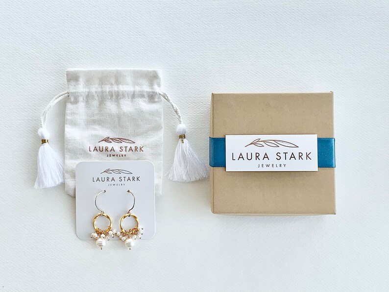 Pu&ograve; includere: Un paio di orecchini a cerchio dorati con piccole perle bianche che pendono dal fondo. Gli orecchini sono esposti su una carta bianca con il testo "LAURA STARK JEWELRY".