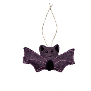 May include: A purple felt bat ornament featuring black bead eyes, a small black oval belly patch, and light purple stitched detailing along the wings and ears. The bat is suspended by a thin, metallic silver string loop for hanging purposes.