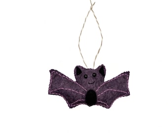 May include: A purple felt bat ornament featuring black bead eyes, a small black oval belly patch, and light purple stitched detailing along the wings and ears. The bat is suspended by a thin, metallic silver string loop for hanging purposes.