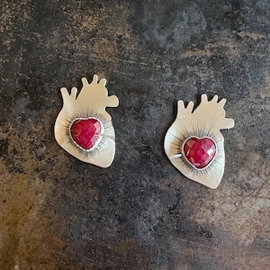 May include: Two silver-toned heart-shaped pendants with a faceted, red gemstone center. The pendants have a detailed anatomical heart design with radiating lines. The background is a dark, textured surface.