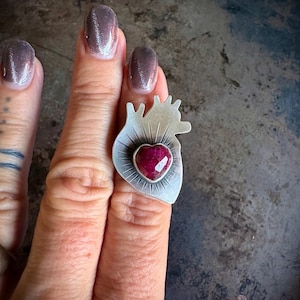 May include: A silver ring featuring a stylized anatomical heart shape with a matte finish and etched radiating lines. A faceted, heart-shaped deep pink gemstone is bezel-set in the center of the silver heart, creating a bold and textured appearance.