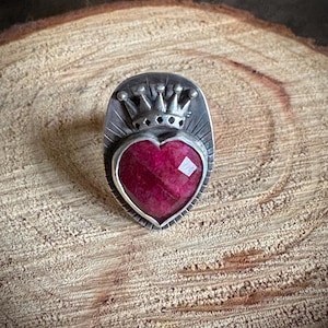 May include: A silver ring featuring a heart-shaped, faceted, deep red gemstone. The ring is topped with a crown design. The ring is set against a textured, wooden background.