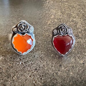 May include: Two silver rings featuring heart-shaped stones set in metal. The left ring has a faceted bright orange stone, while the right ring has a smooth deep red stone. Both rings are topped with a decorative silver rose and leaf motif.
