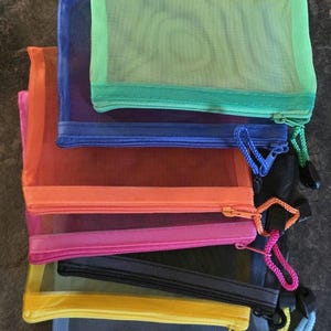 May include: A stack of colorful mesh zipper pouches in shades of green, blue, orange, pink, yellow, and gray. Each pouch has a matching zipper and a small loop for carrying. The pouches are designed for organizing and storing items.