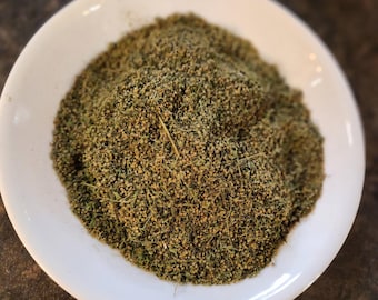 May include: A white ceramic plate filled with a mound of dried, green herbs. The herbs have a fine, granular texture and a mix of light and dark green hues. A few thin, light-colored stems are visible within the pile.