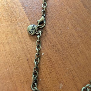 May include: Close-up of an antique brass-coloured necklace chain with a lobster clasp. A small, heart-shaped charm with the word "LOVE" is attached near the clasp. The chain is on a wooden surface.