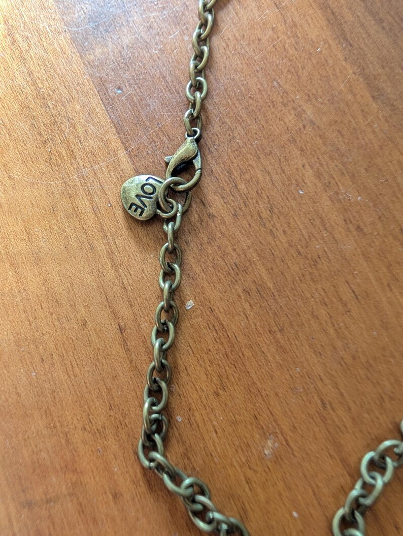 May include: Close-up of an antique brass-coloured necklace chain with a lobster clasp. A small, heart-shaped charm with the word "LOVE" is attached near the clasp. The chain is on a wooden surface.