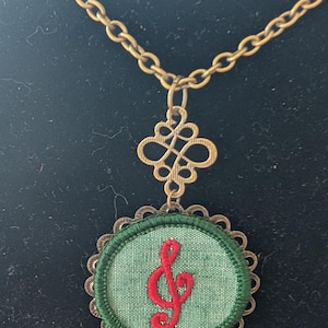 May include: A brass-coloured necklace with a green fabric pendant embroidered with a red treble clef. The pendant has a scalloped edge and a small key charm. The chain and decorative elements have an antique finish.