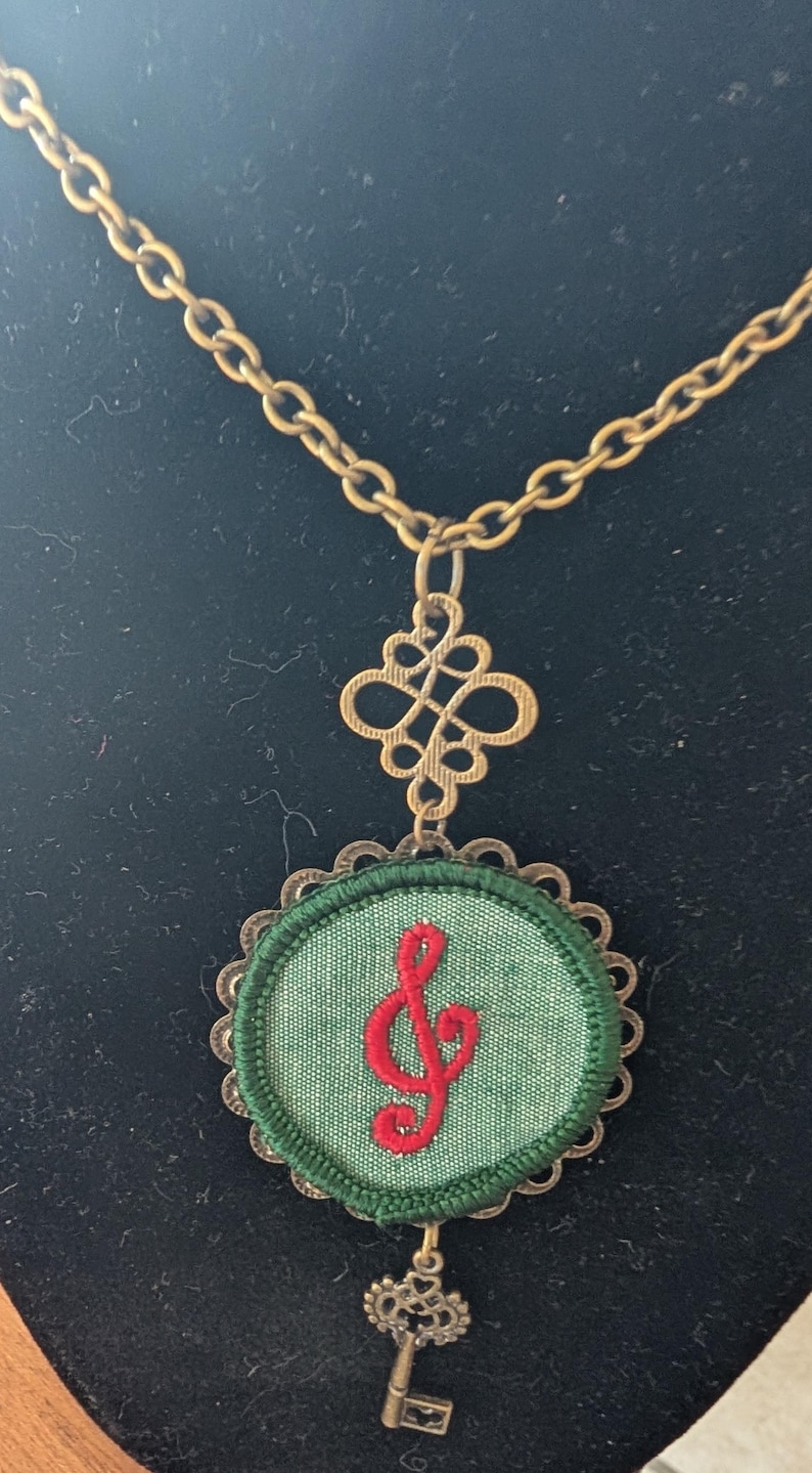 May include: A brass-coloured necklace with a green fabric pendant embroidered with a red treble clef. The pendant has a scalloped edge and a small key charm. The chain and decorative elements have an antique finish.