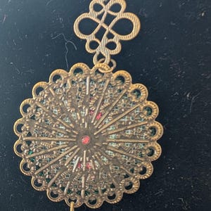 May include: An antique-style brass necklace with an elaborate, filigree pendant. The pendant has a central red stone and a small key charm. The chain and other elements are brass-toned, giving it an antique appearance. The necklace is a decorative accessory.