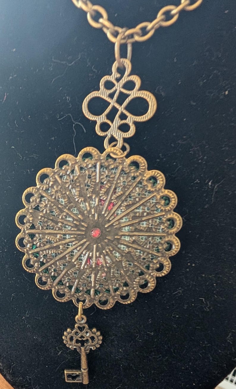 May include: An antique-style brass necklace with an elaborate, filigree pendant. The pendant has a central red stone and a small key charm. The chain and other elements are brass-toned, giving it an antique appearance. The necklace is a decorative accessory.