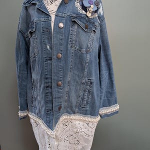 Könnte beinhalten: Hellblaue Jeansjacke mit weißer Spitze und Perlenbesatz. Die Jacke hat eine dekorative blaue Blume mit lila Mitte und weißen Spitzendetails. Die Jacke hat silberne Knöpfe und wird auf einer Schaufensterpuppe präsentiert.