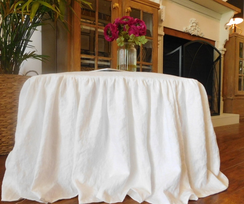 Puede incluir: Falda de mesa de lino blanco con un borde fruncido. La falda est&aacute; drapeada sobre una mesa redonda y est&aacute; en una habitaci&oacute;n con una chimenea y una librer&iacute;a.