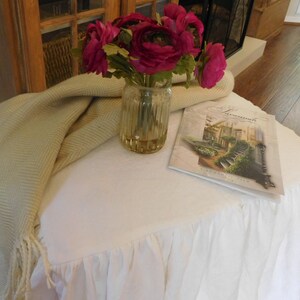 Puede incluir: Una mesa blanca con una falda con volantes, una manta de tiro beige, un jarr&oacute;n de flores rosas y un libro titulado "Landscapes" en la parte superior.