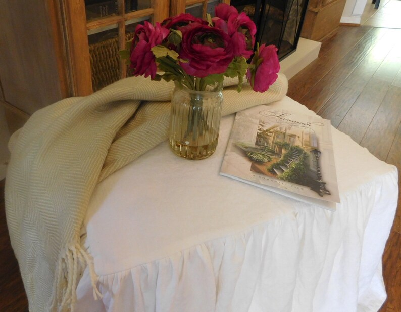 Puede incluir: Una mesa blanca con una falda con volantes, una manta de tiro beige, un jarr&oacute;n de flores rosas y un libro titulado "Landscapes" en la parte superior.