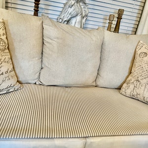 May include: A white couch with beige cushions and a striped throw. The couch is in front of a window with white blinds. There is a white horse head sculpture on the back of the couch.