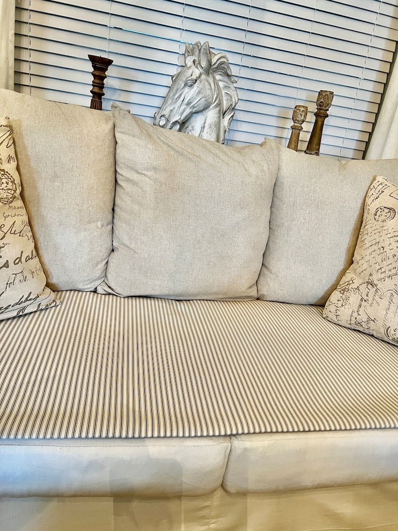 May include: A white couch with beige cushions and a striped throw. The couch is in front of a window with white blinds. There is a white horse head sculpture on the back of the couch.