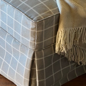 May include: A grey and white checked fabric ottoman with a white fringe throw blanket draped over the top.