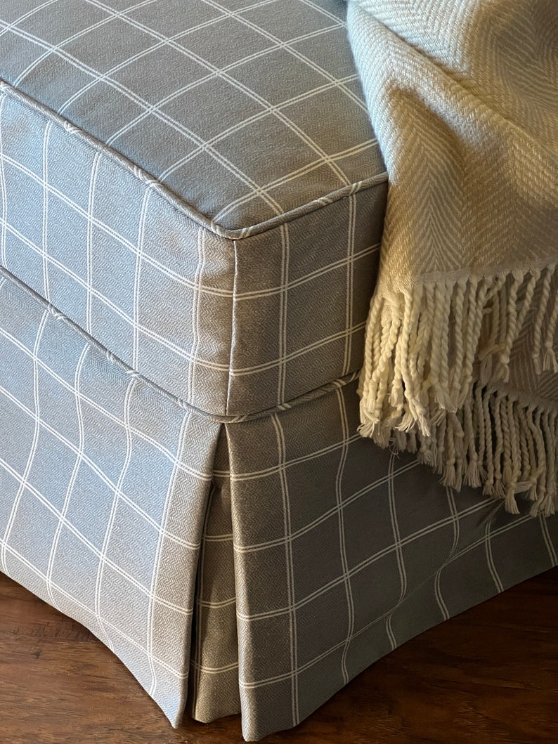 May include: A grey and white checked fabric ottoman with a white fringe throw blanket draped over the top.