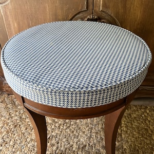 May include: A round, upholstered stool with a dark wood frame. The seat features a blue and white houndstooth pattern. The stool has curved legs and is set on a woven rug. A wooden cabinet is in the background.