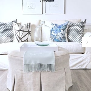 May include: A round ottoman with a light beige pleated skirt, topped with a light blue fringed throw. A white tray holds a clear glass bottle. Decorative pillows in blue and white patterns sit on a white sofa.