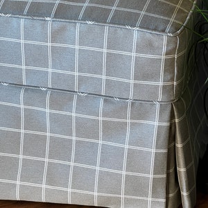May include: A grey and white checked ottoman with a pleated skirt. The ottoman is made of a soft, textured fabric.