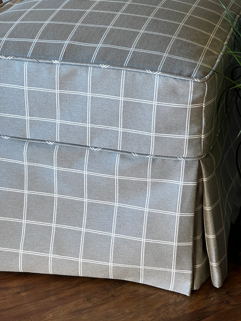 May include: A grey and white checked ottoman with a pleated skirt. The ottoman is made of a soft, textured fabric.