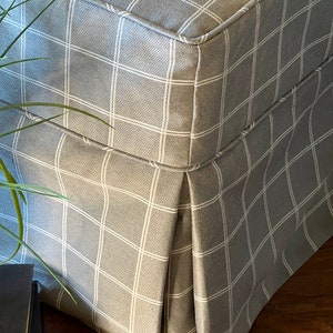 May include: A grey and white checked ottoman with a pleated skirt. The ottoman is made of a soft, textured fabric.