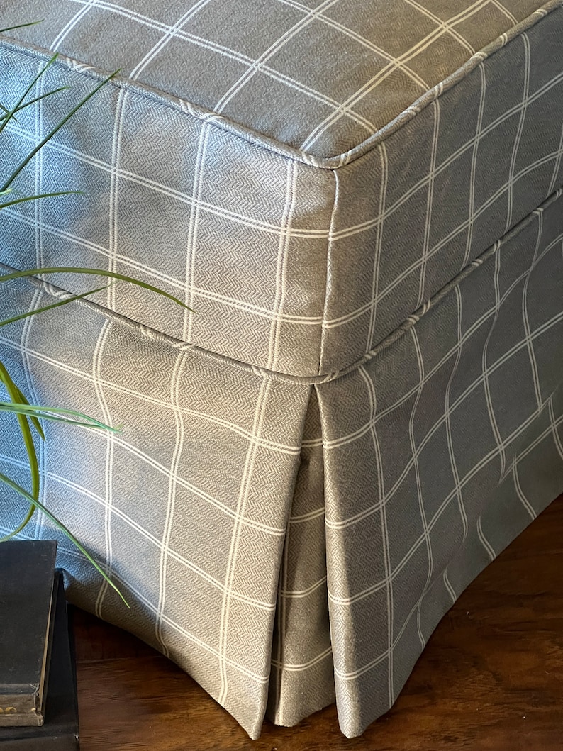 May include: A grey and white checked ottoman with a pleated skirt. The ottoman is made of a soft, textured fabric.