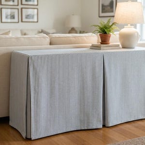 May include: A rectangular console table covered by a floor-length fabric slipcover featuring a blue and white pinstripe pattern. The slipcover has two distinct box pleats along the front panel, creating a structured and tailored appearance.