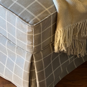 May include: A gray and white checkered fabric ottoman with a pleated skirt and a white throw blanket with a fringe.