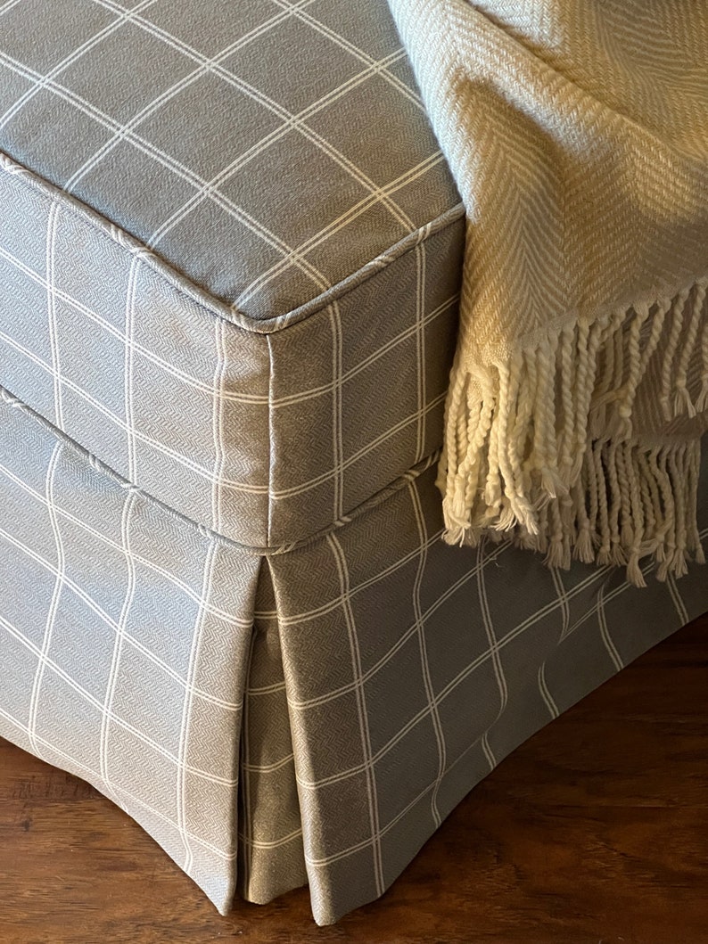 May include: A grey and white checked fabric ottoman with a pleated skirt and a white throw blanket with a fringe.
