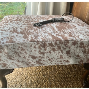 May include: A brown and white cowhide ottoman with four wooden legs. A decorative key with a ring is on top of the ottoman.