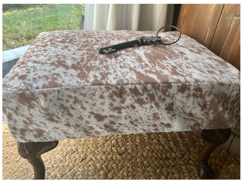 May include: A brown and white cowhide ottoman with four wooden legs. A decorative key with a ring is on top of the ottoman.