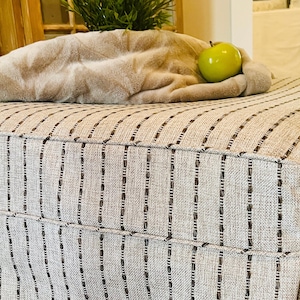 May include: A striped ottoman with a brown and white fabric cover. A soft, beige throw blanket and a green apple are on top of the ottoman.