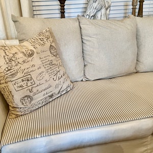 May include: A white couch with a striped throw and three pillows. The pillows are beige and have a brown and white pattern with handwritten text. The couch is in a living room setting.