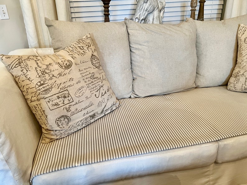May include: A white couch with a striped throw and three pillows. The pillows are beige and have a brown and white pattern with handwritten text. The couch is in a living room setting.