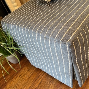 May include: A blue upholstered ottoman with vertical white stripes. The ottoman has a pleated skirt and sits on a wooden floor. A small potted plant with long green leaves is visible on the left.