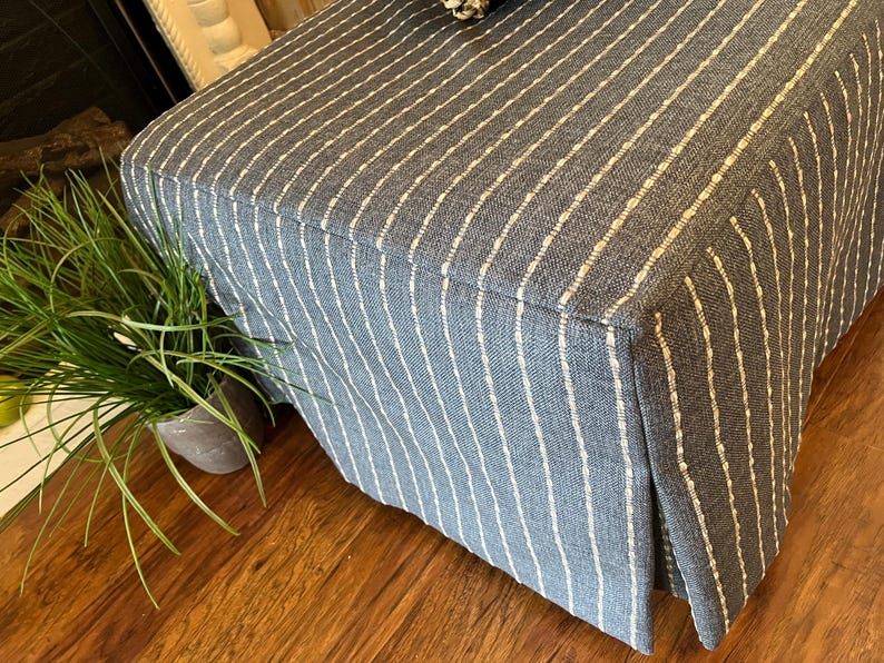 May include: A blue upholstered ottoman with vertical white stripes. The ottoman has a pleated skirt and sits on a wooden floor. A small potted plant with long green leaves is visible on the left.