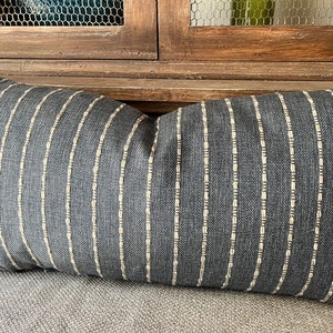 May include: A rectangular decorative pillow with a dark gray fabric and vertical cream-colored stripes. The pillow is placed on a gray surface, with a wooden cabinet in the background.